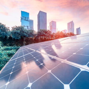 In the foreground there is zoomed in view of a solar panel in a natural background of green grass and trees. In the background there is a city skyline with a sunrise background.