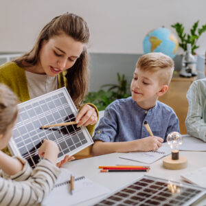 A teacher demonstrates a solar panel to elementary students gathered around a table taking notes, with a globe visible in the bright classroom behind them.